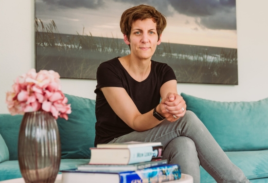 Bohana-Gründerin Anne Kriesel schaut lächelnd in die Kamera. Sie sitzt auf einem grünen Sofa mit einer Blumenvase und Büchern im Vordergrund.